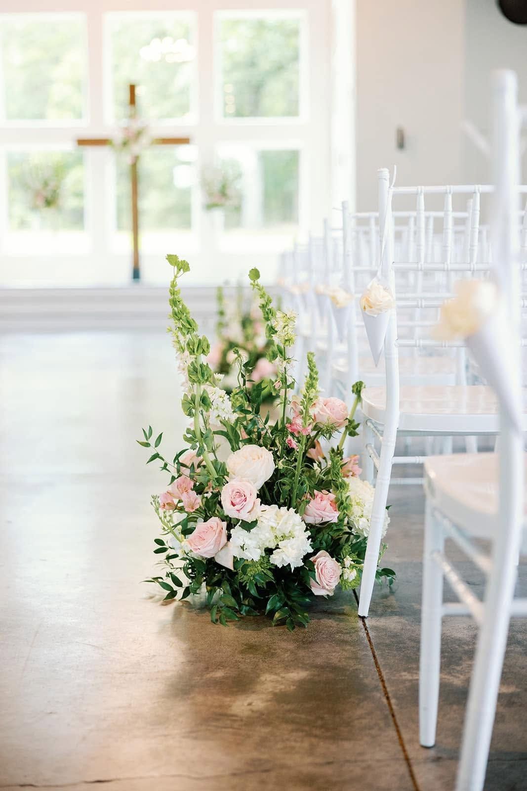 White chairs line a bright aisle with a pastel pink-and-green floral arrangement beside them