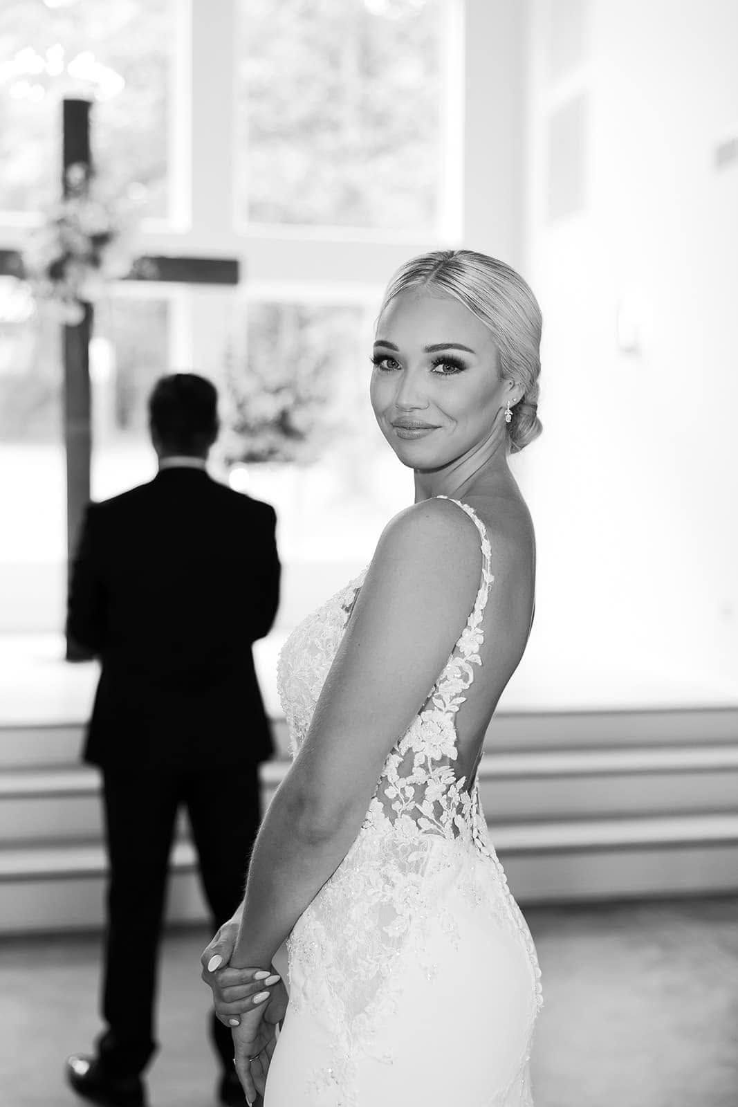 Smiling bride in a white lace gown posing indoors, with a man blurred in the background