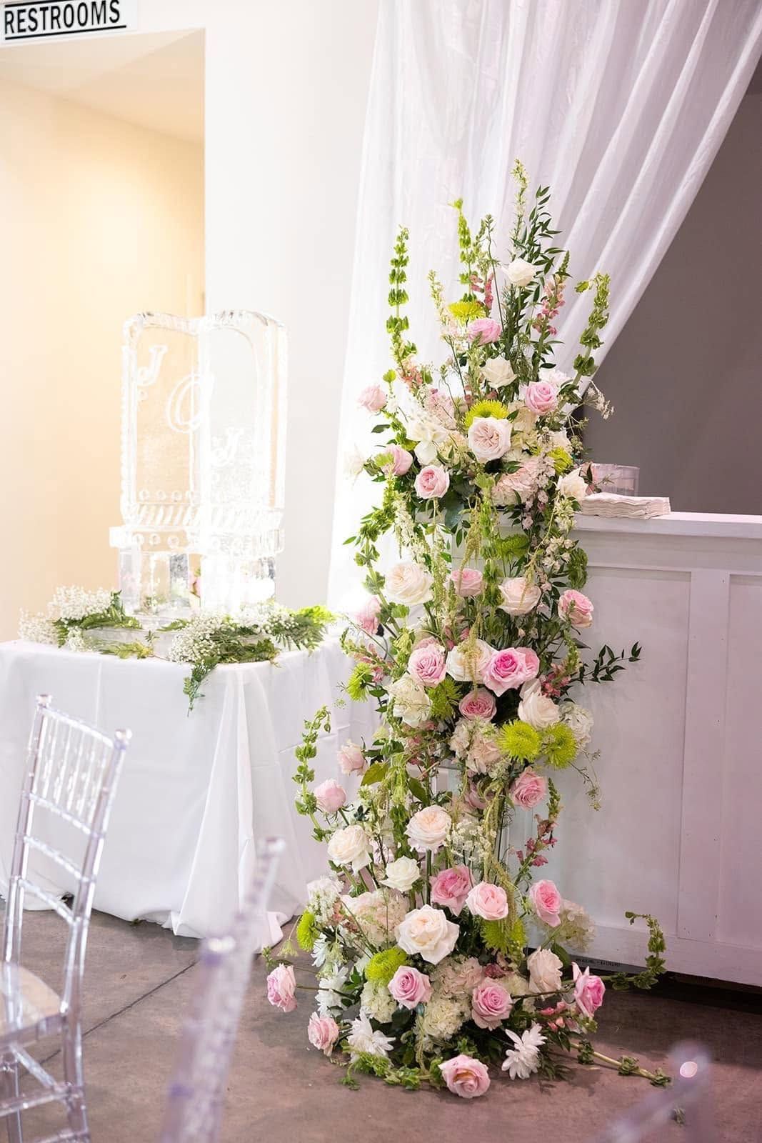 Tall pink and white floral arrangement beside a draped white wedding table in a bright venue