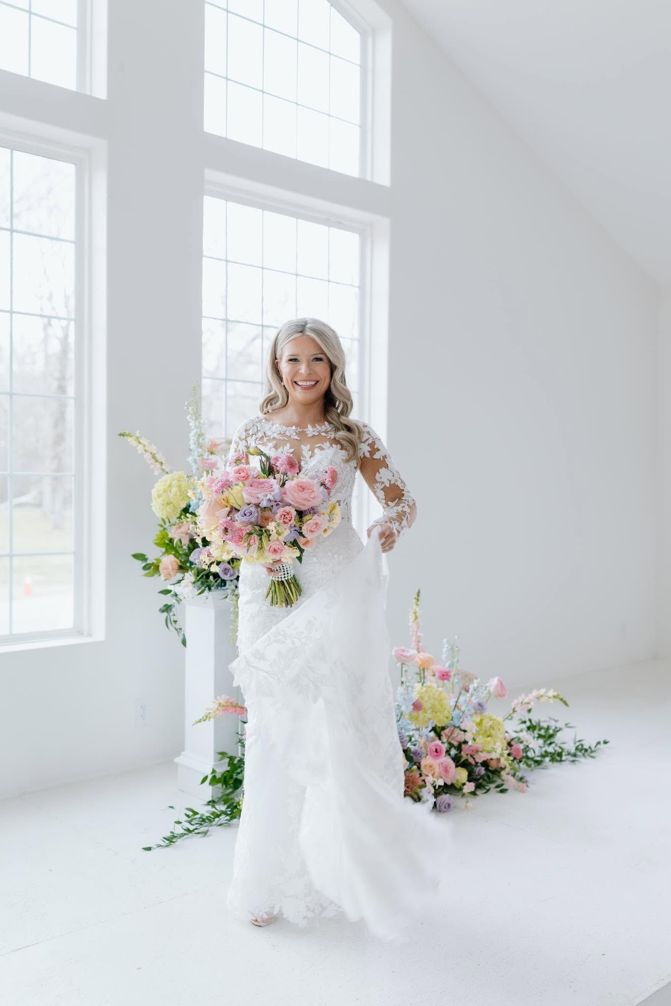 Bride in white lace gown holding a pastel bouquet in a bright white room with large windows