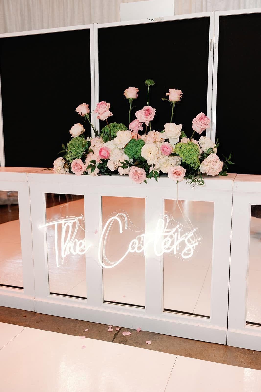 Neon sign on white railing beneath pink floral arrangements against black panels.
