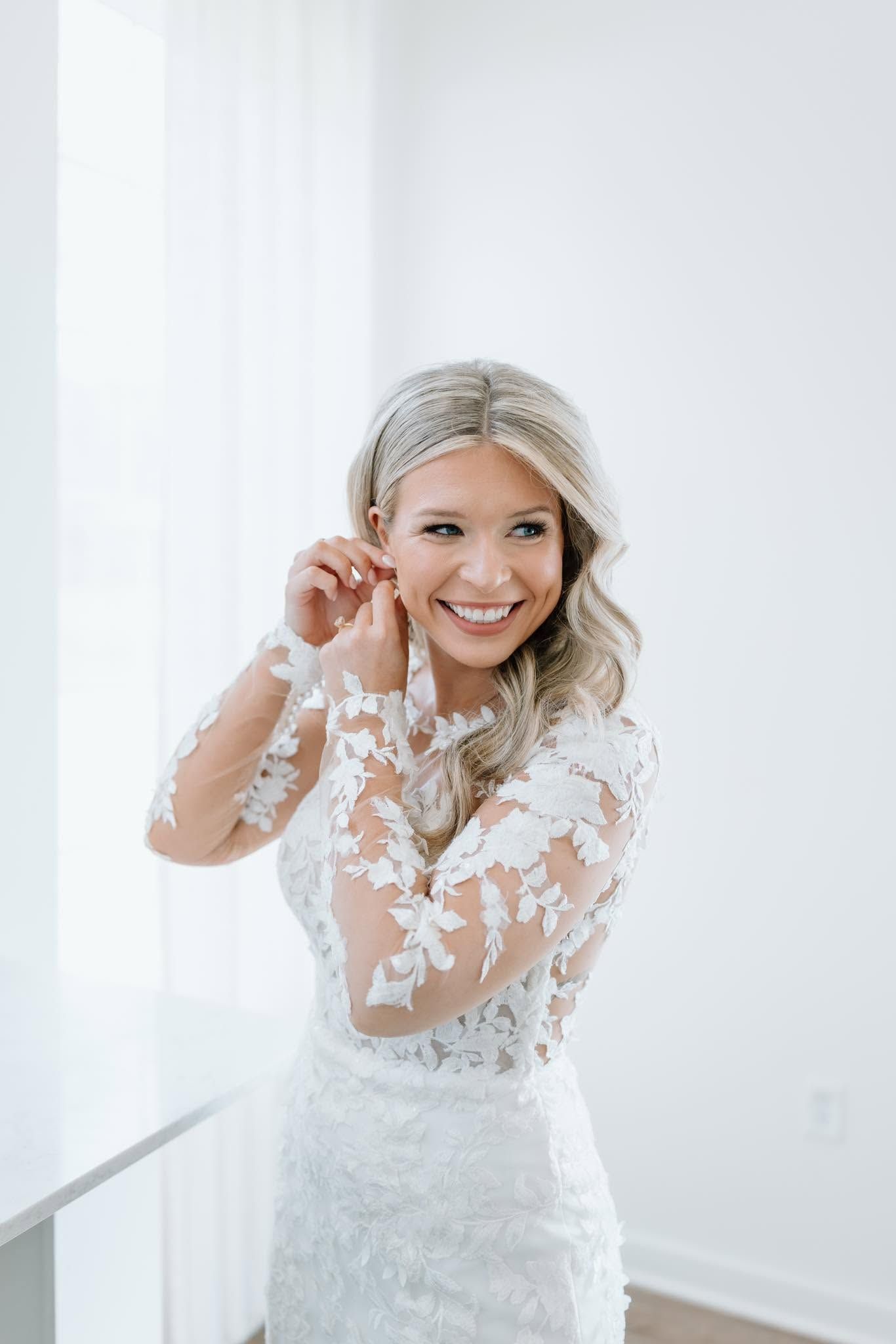 Woman in a white lace dress smiling while adjusting her earring in a bright room.