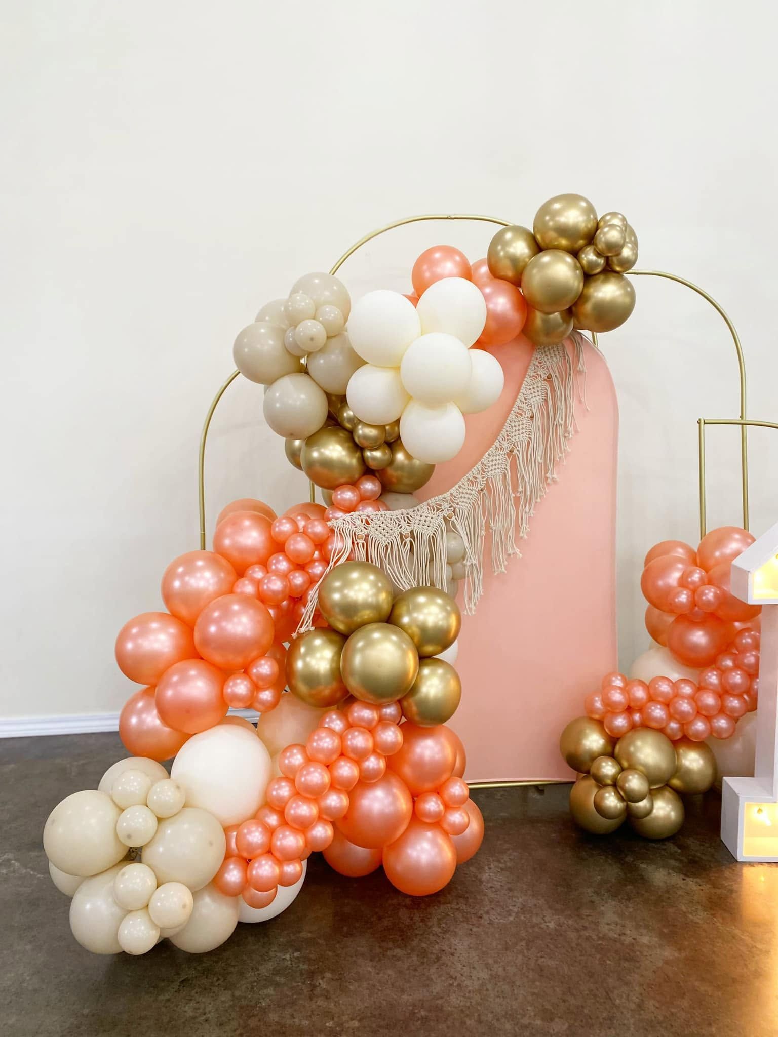 Peach, ivory, and gold balloon arch framing a pink panel and tassel backdrop on a wood floor