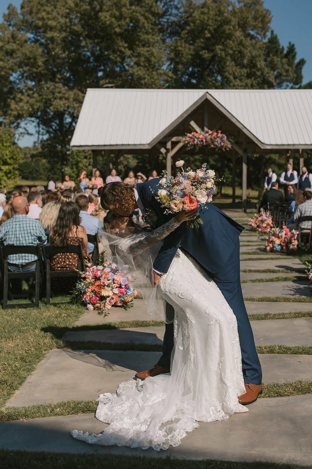 Wedding couple embracing outdoors at an aisle-lined ceremony, with floral arrangements and a pavilion in the background
