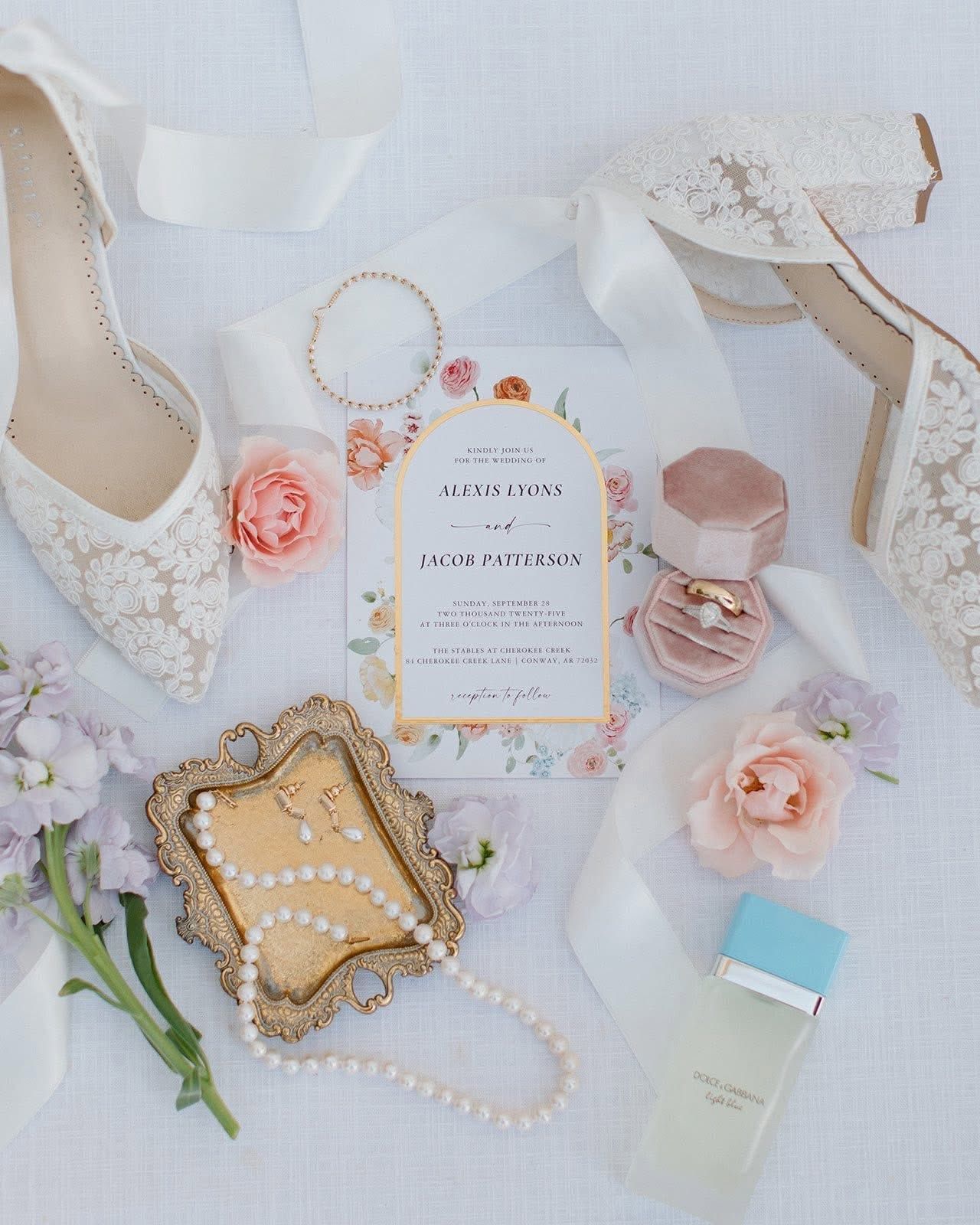 Elegant bridal flat lay with white lace lingerie, jewelry, flowers, perfume, and invitation on soft white fabric