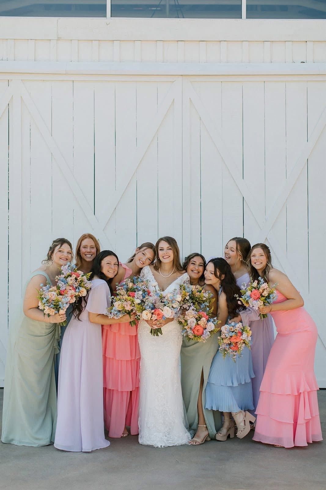 A group of women in pastel dresses and a white gown holding bouquets, posing in front of white barn doors.