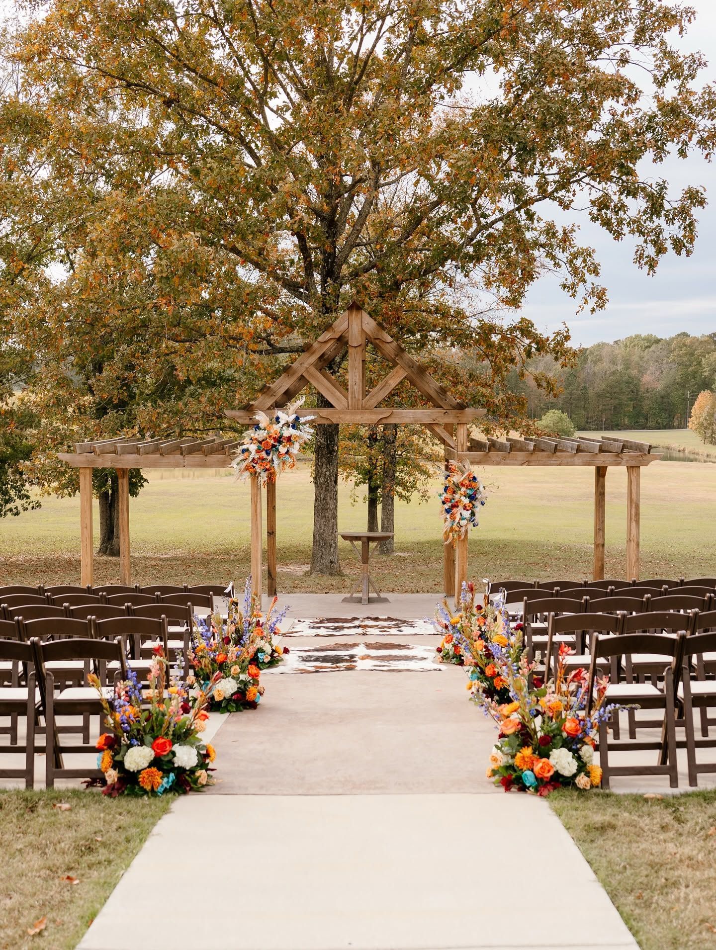 Outdoor wedding aisle with floral arrangements, wooden chairs, and a rustic arch under autumn trees
