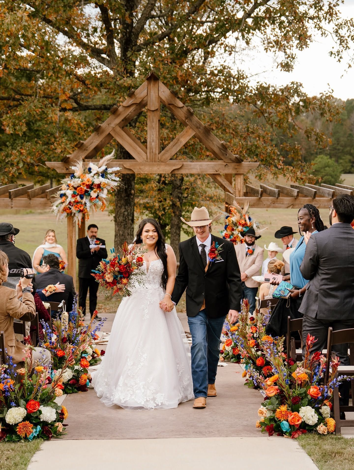 Bride and groom walking down a flower-lined outdoor wedding aisle under a wooden arch