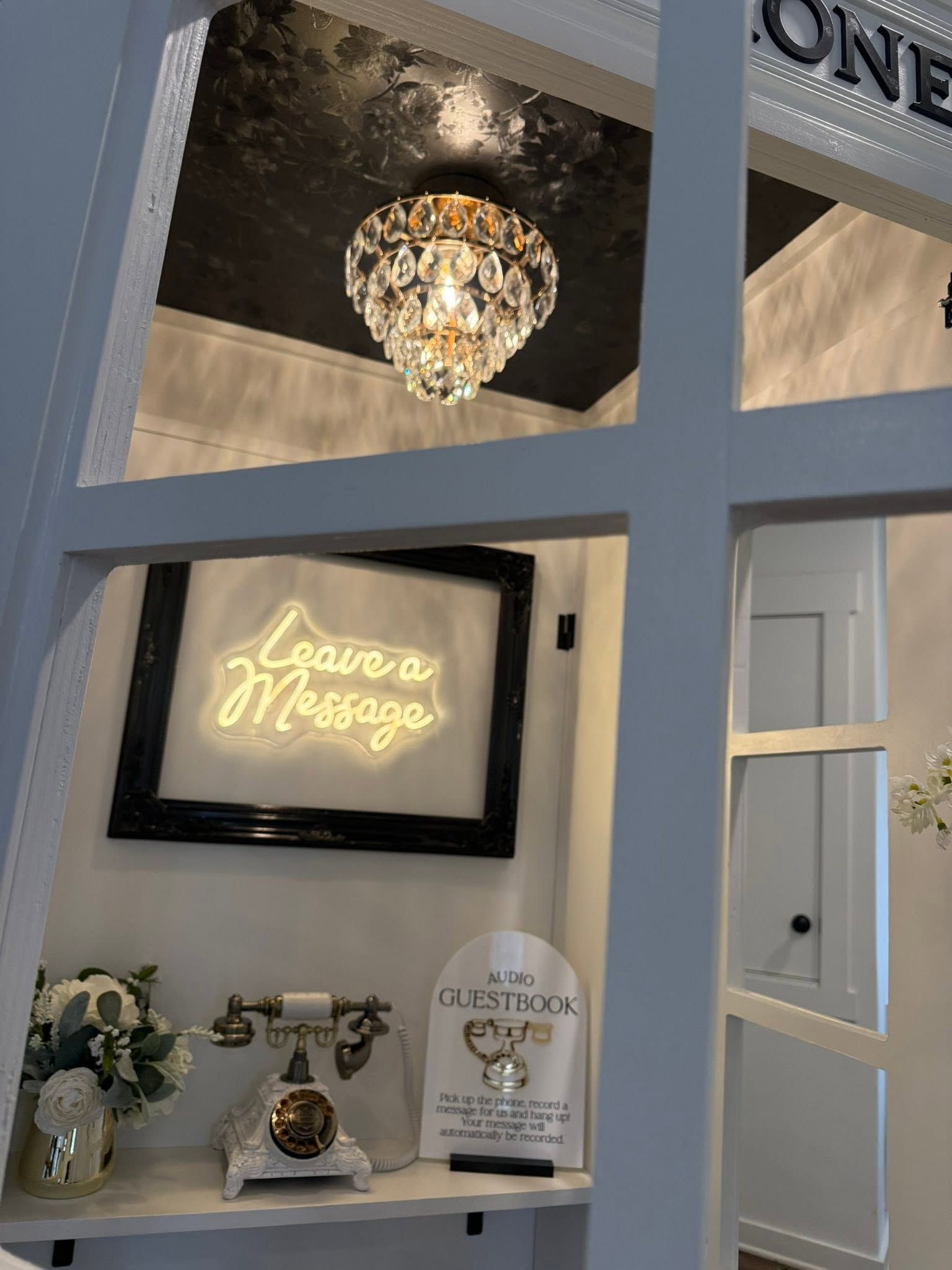 Neon “Love Message” sign above a decorated mantel with flowers, clock, and chandelier in a white room