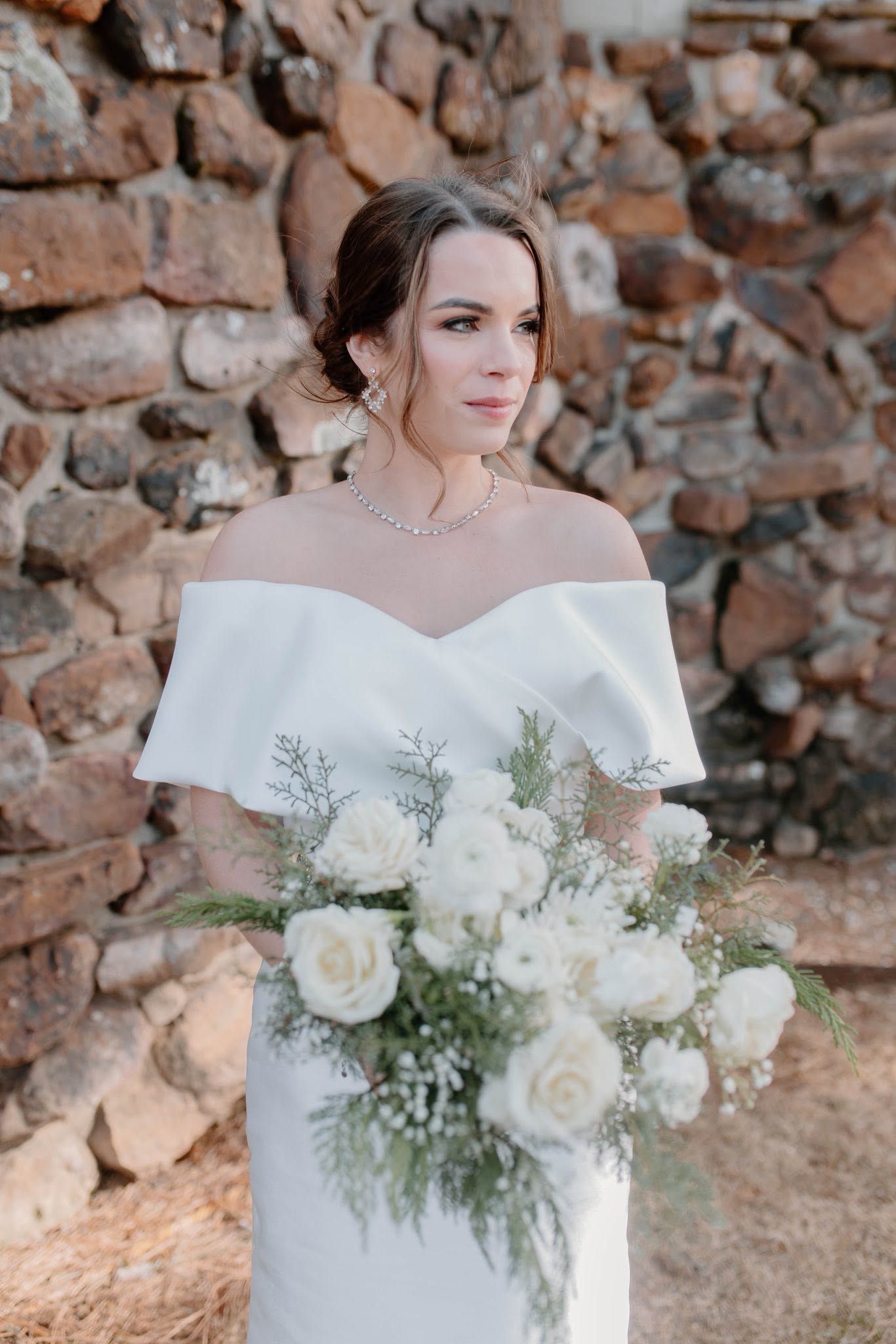 Bride in a white off-shoulder dress holding a large white bouquet in front of a stone wall