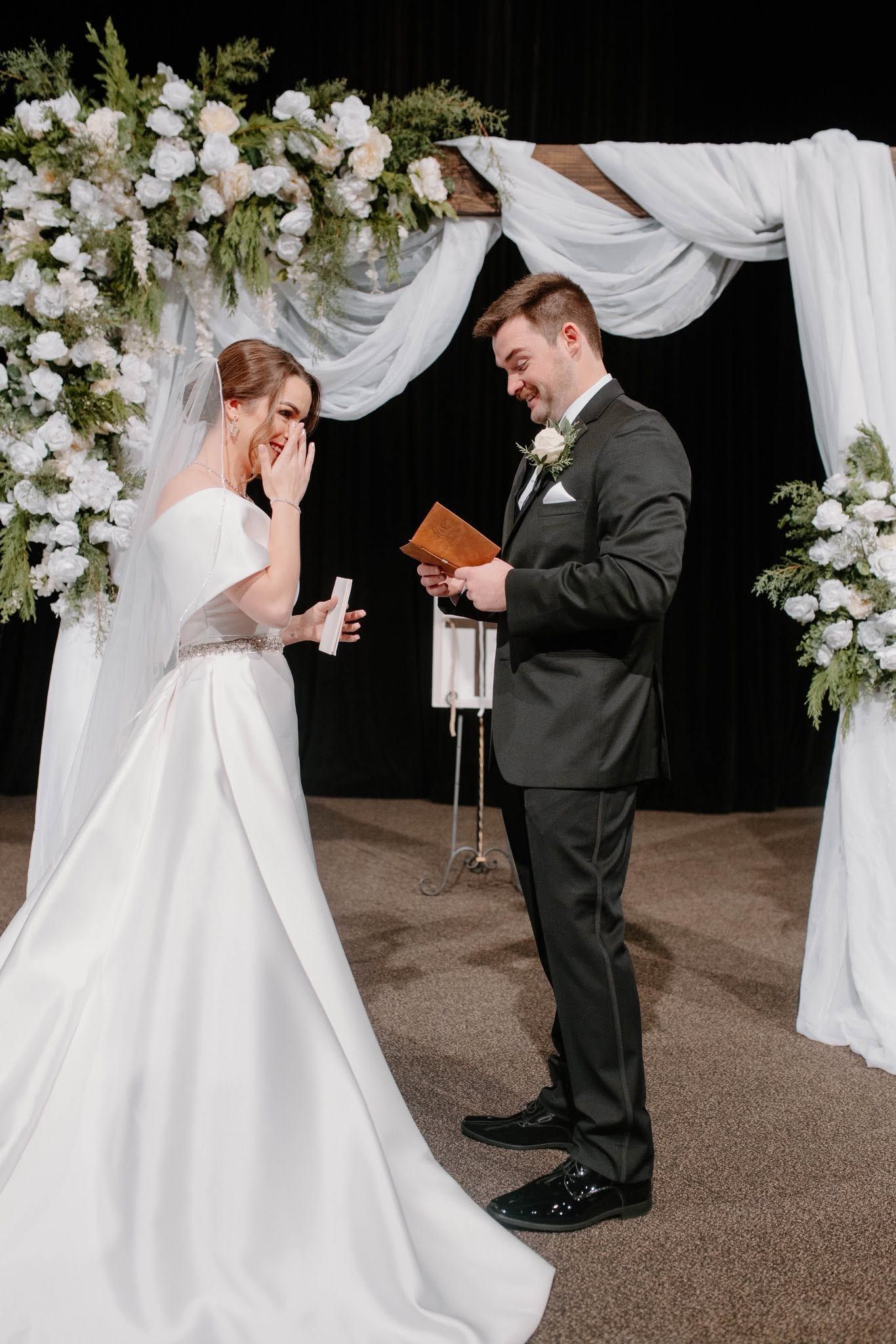 Bride and groom exchanging vows under a white floral arch at an indoor wedding ceremony