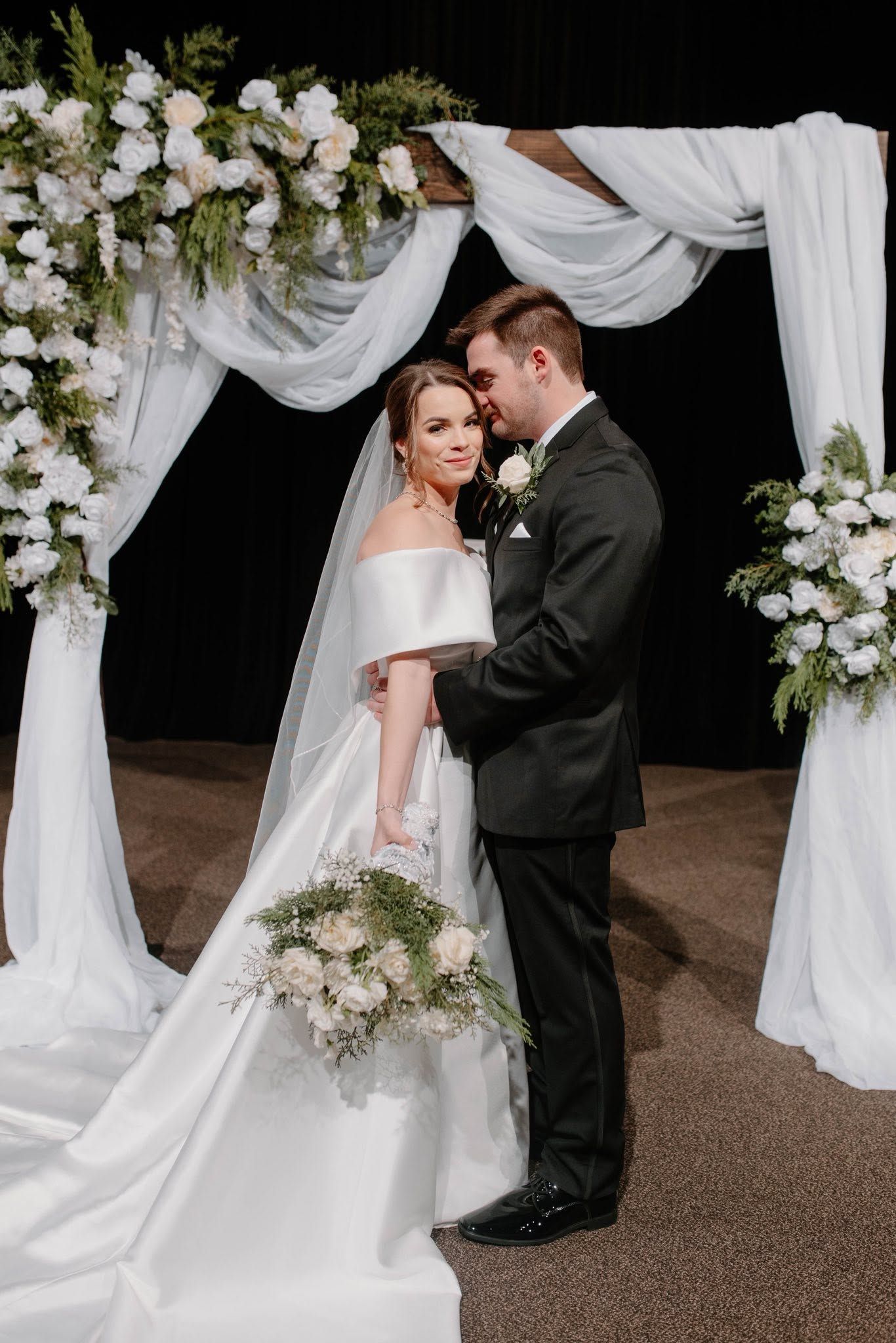 Bride and groom in white and black attire stand beneath a floral wedding arch at night.