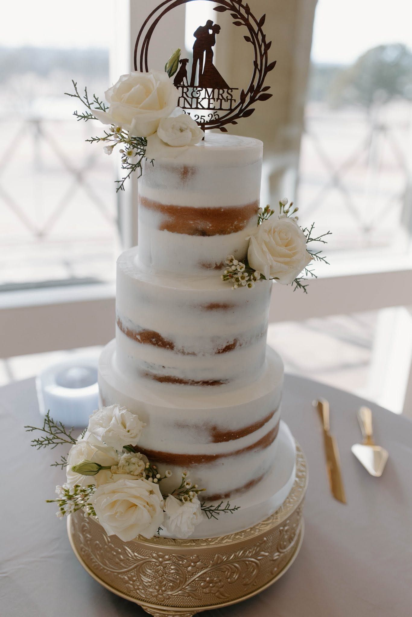 White tiered wedding cake with white flowers and gold accents on a table near a window