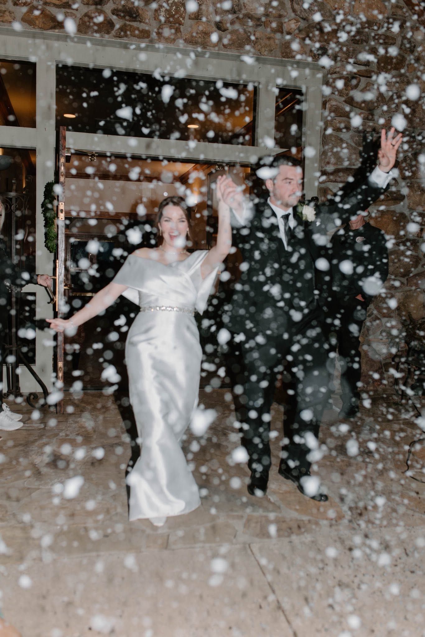 Newlyweds in wedding attire walking through falling confetti outside a building