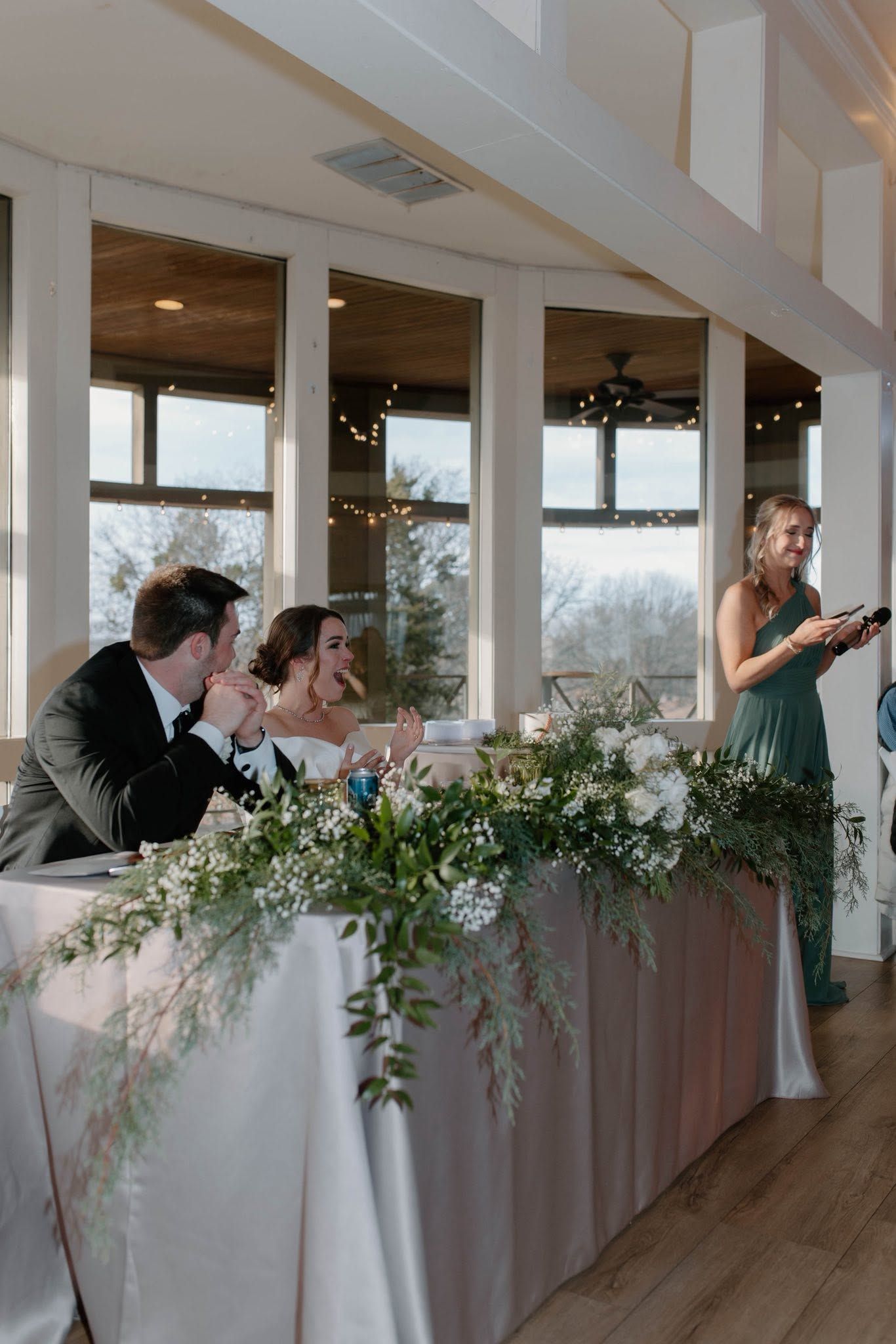 Wedding reception with seated guests and a woman speaking beside a white-draped head table with greenery