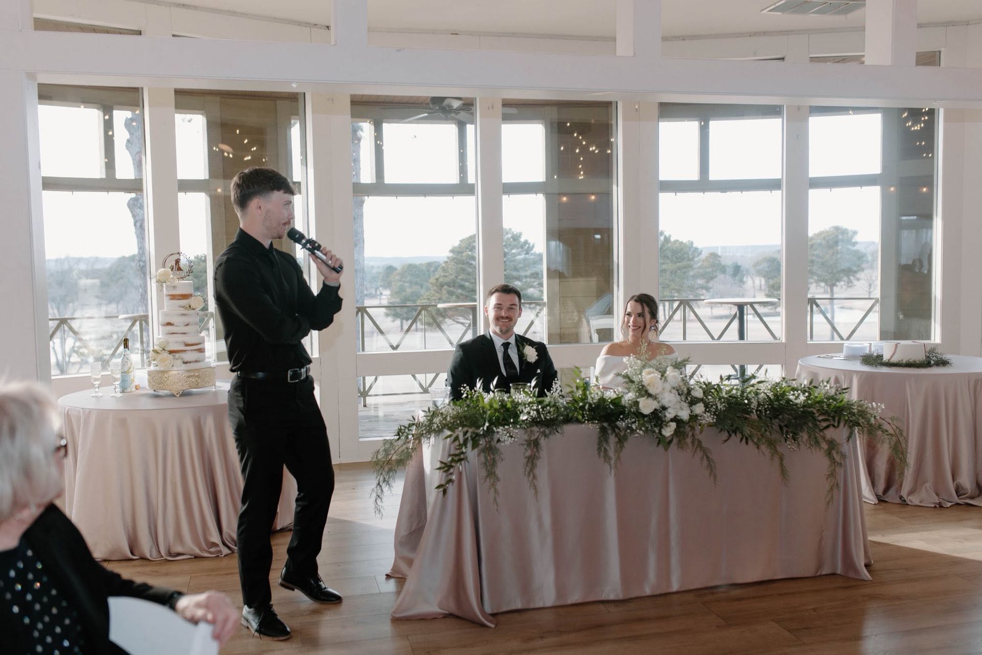 Speaker at a wedding reception addresses seated guests beside a decorated head table in a bright room