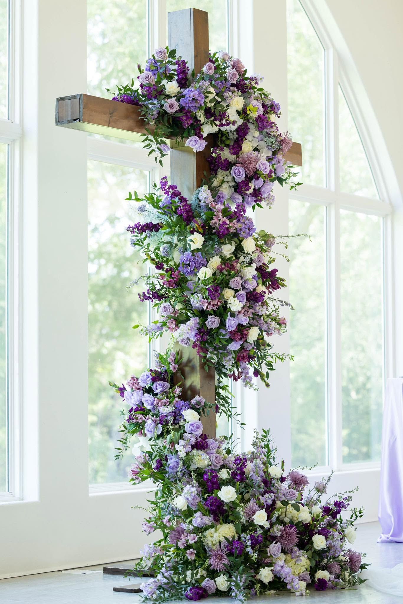 Purple and white flower arrangement cascading from a cross-shaped stand in a bright windowed room