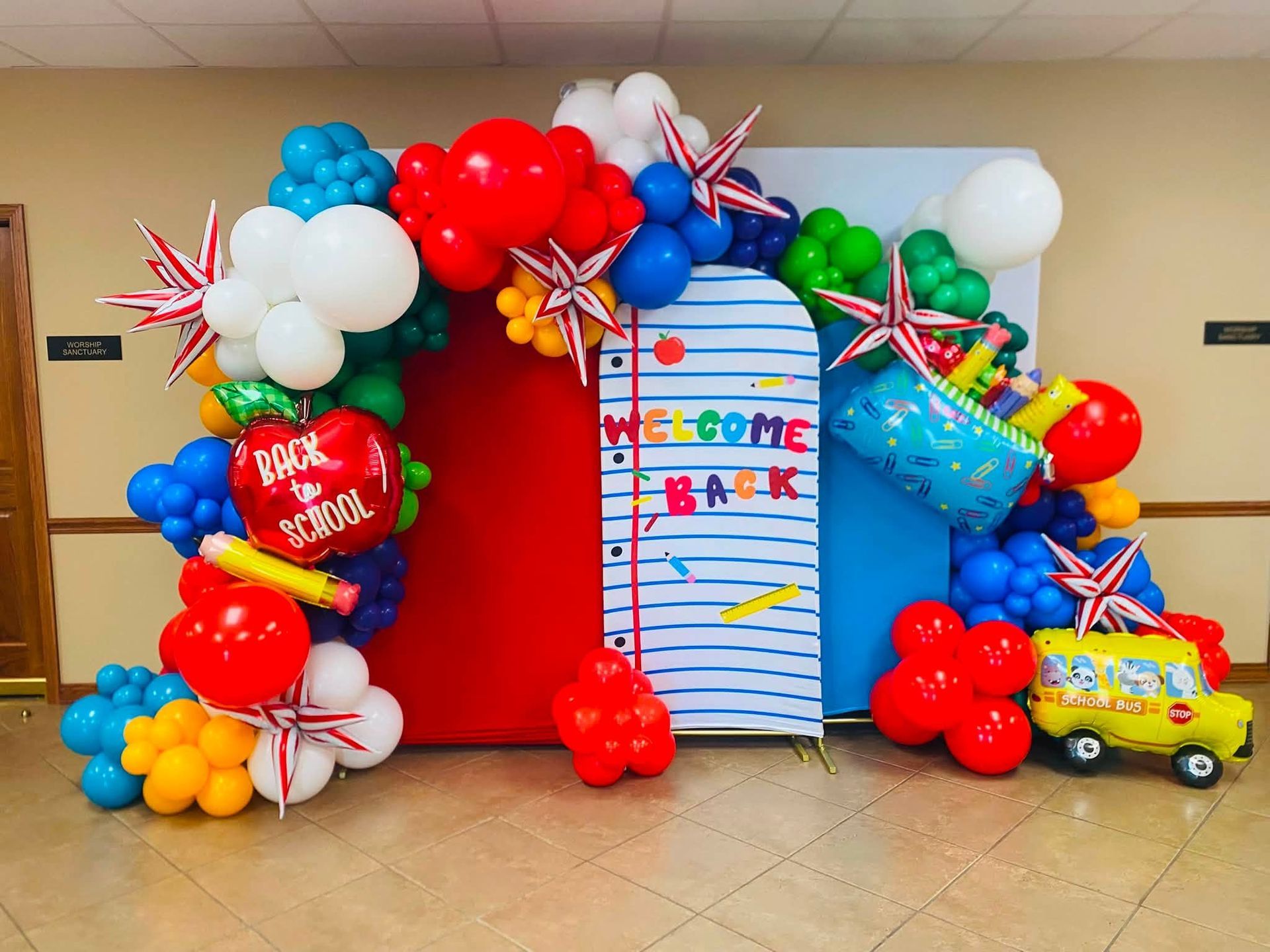 Colorful balloon arch framing a red backdrop and striped welcome sign in a hallway; toy construction truck at right.