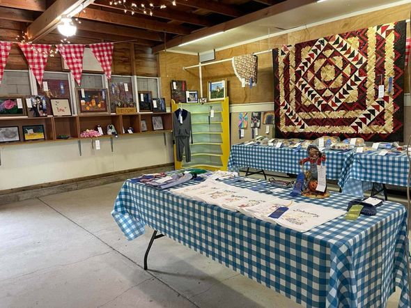 Quilt display in a rustic hall with checkered tablecloths, hanging quilts, and craft items on shelves