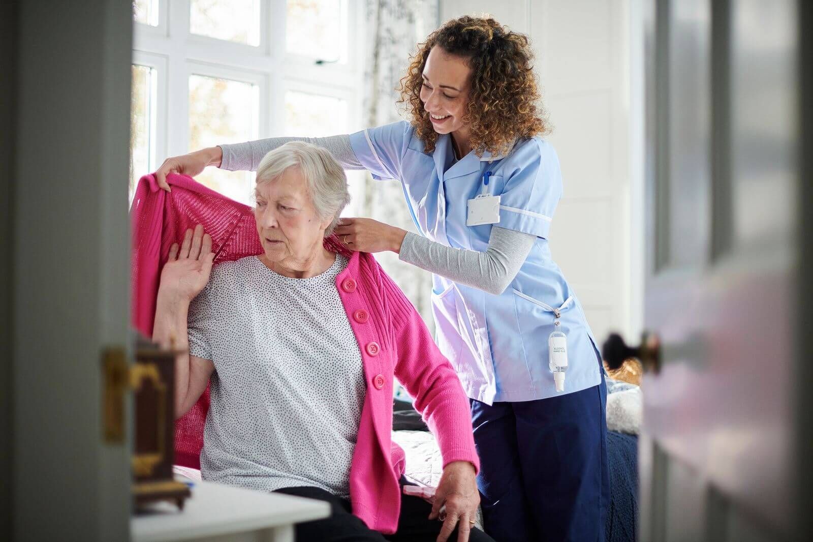 Caregiver helping an older woman into a bright pink cardigan in a sunlit room