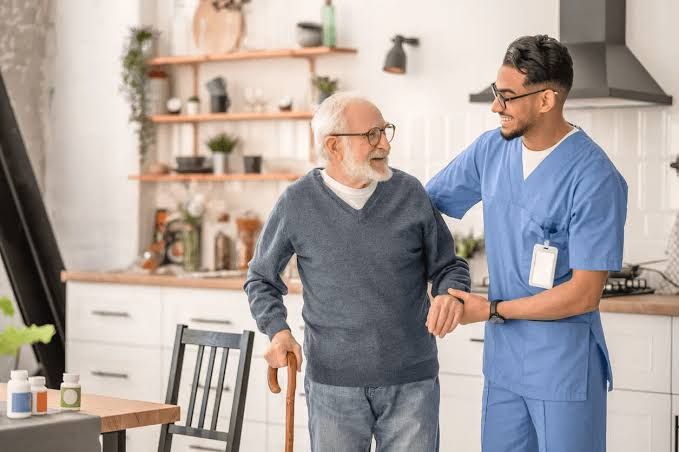 Caregiver in blue scrubs helping an elderly man walk in a bright home kitchen