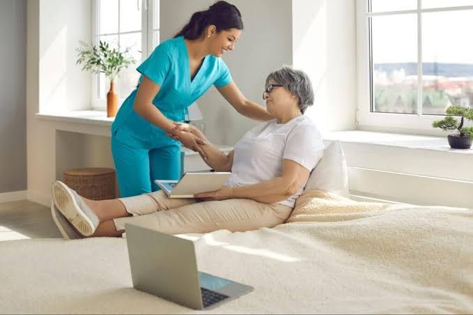 Caregiver helping an older adult sit up on a bed with a laptop nearby in a bright bedroom