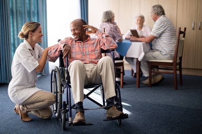 Nurse kneels beside a man in a wheelchair, talking with him in a care facility lounge.