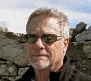 Man wearing sunglasses, against a stone wall background, looking toward the viewer.