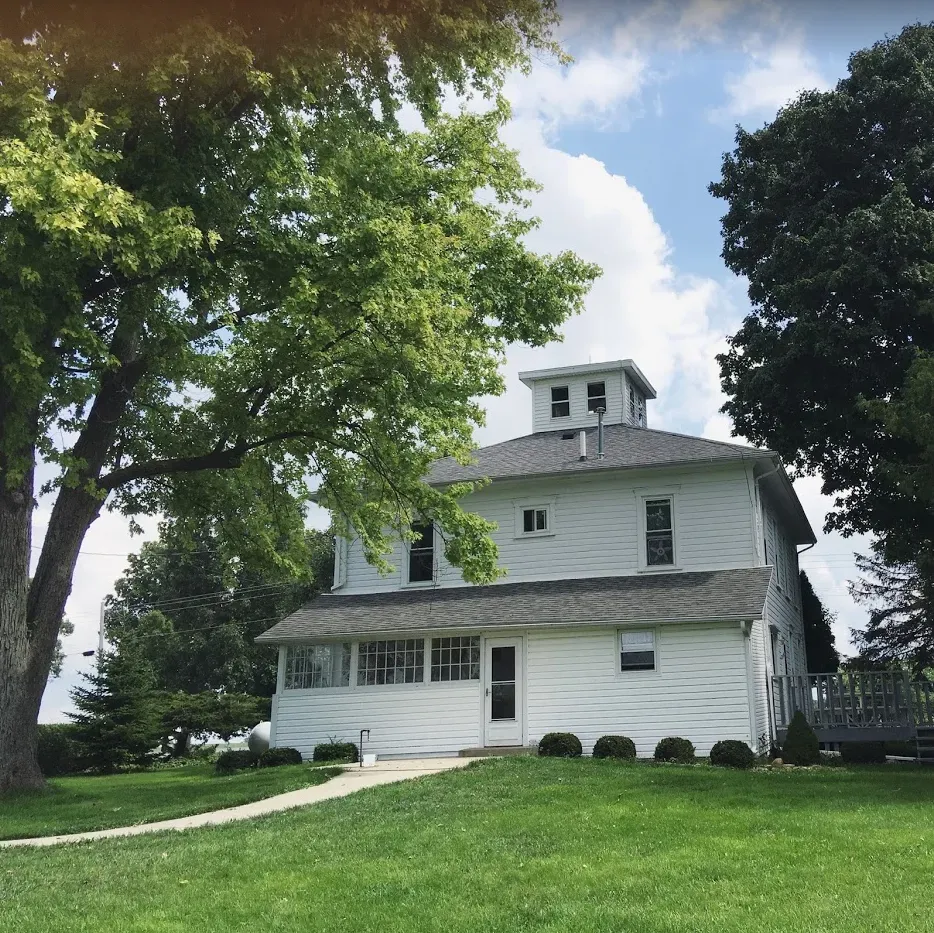 White two-story house with a cupola, porch, and lawn, framed by large trees on a sunny day.
