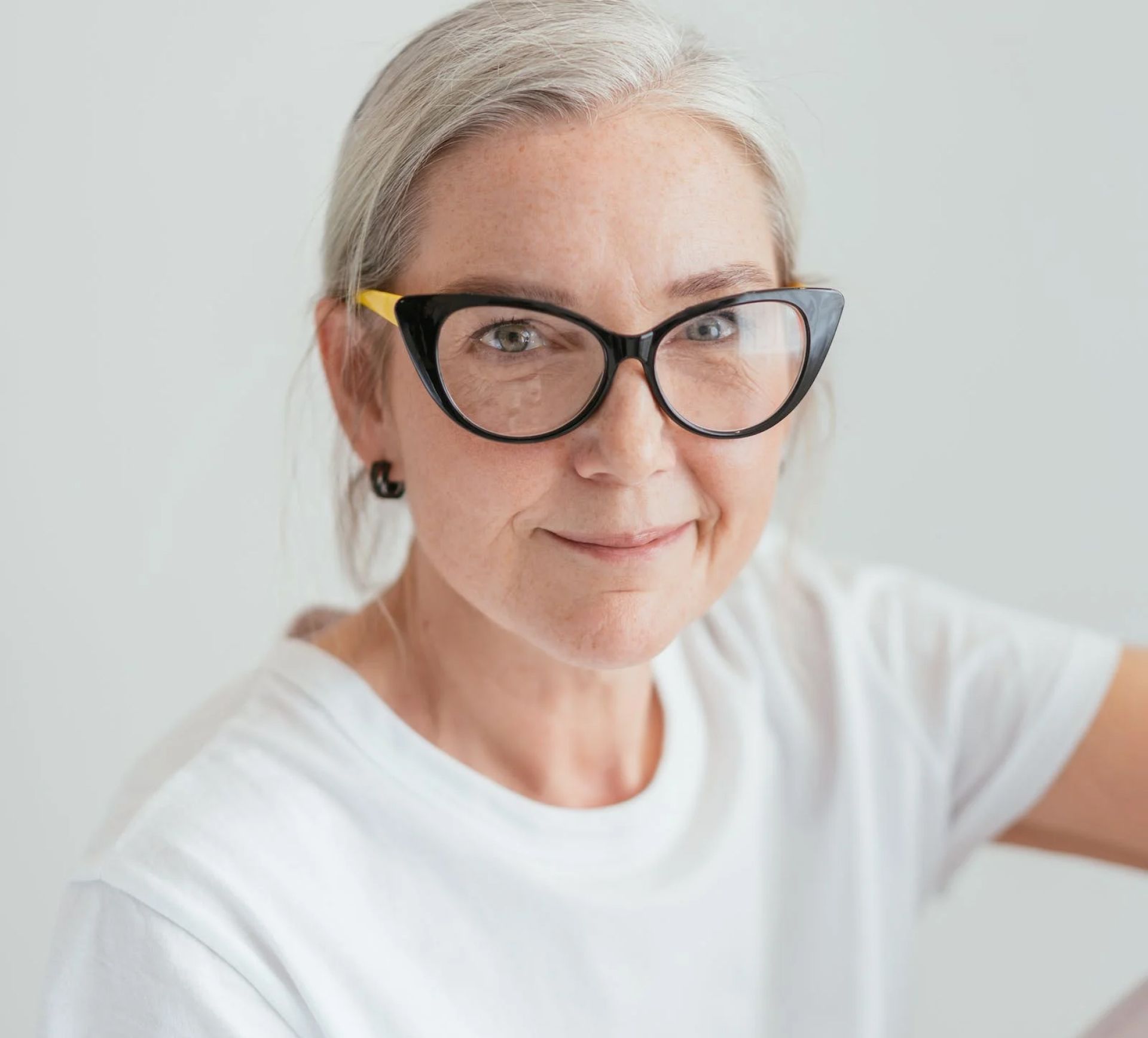 Woman with gray hair wearing black cat-eye glasses and a white t-shirt, smiling at the camera.
