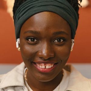 Woman with dark skin and a green headband smiles, wearing earbuds and a collared shirt.