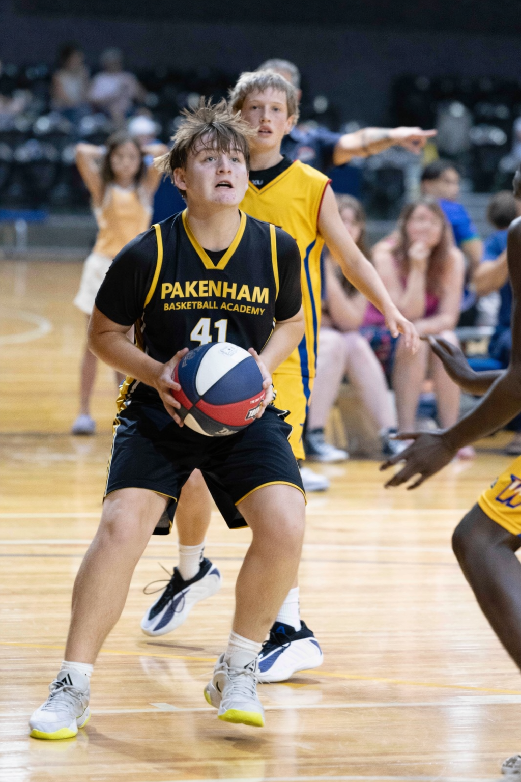 Basketball player in black jersey dribbles on court, with teammate behind and spectators in background