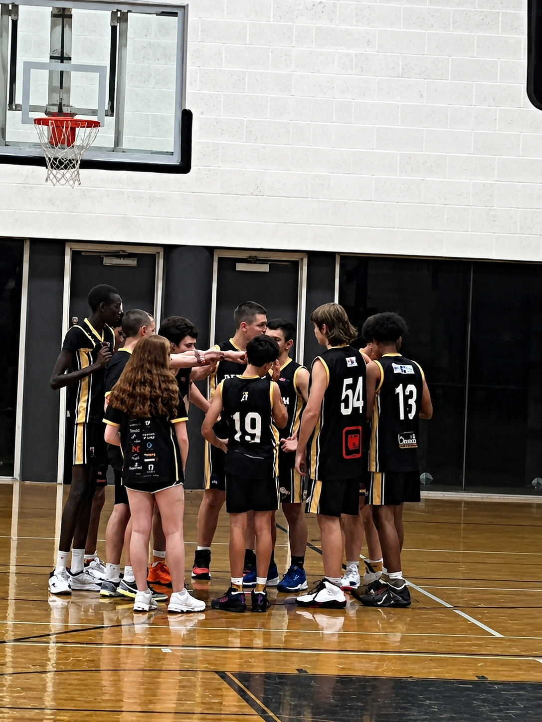 Basketball team huddled under the hoop on an indoor court, black jerseys with red numbers.