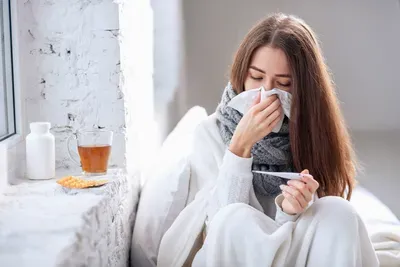 A woman is sitting on a window sill blowing her nose and holding a thermometer.