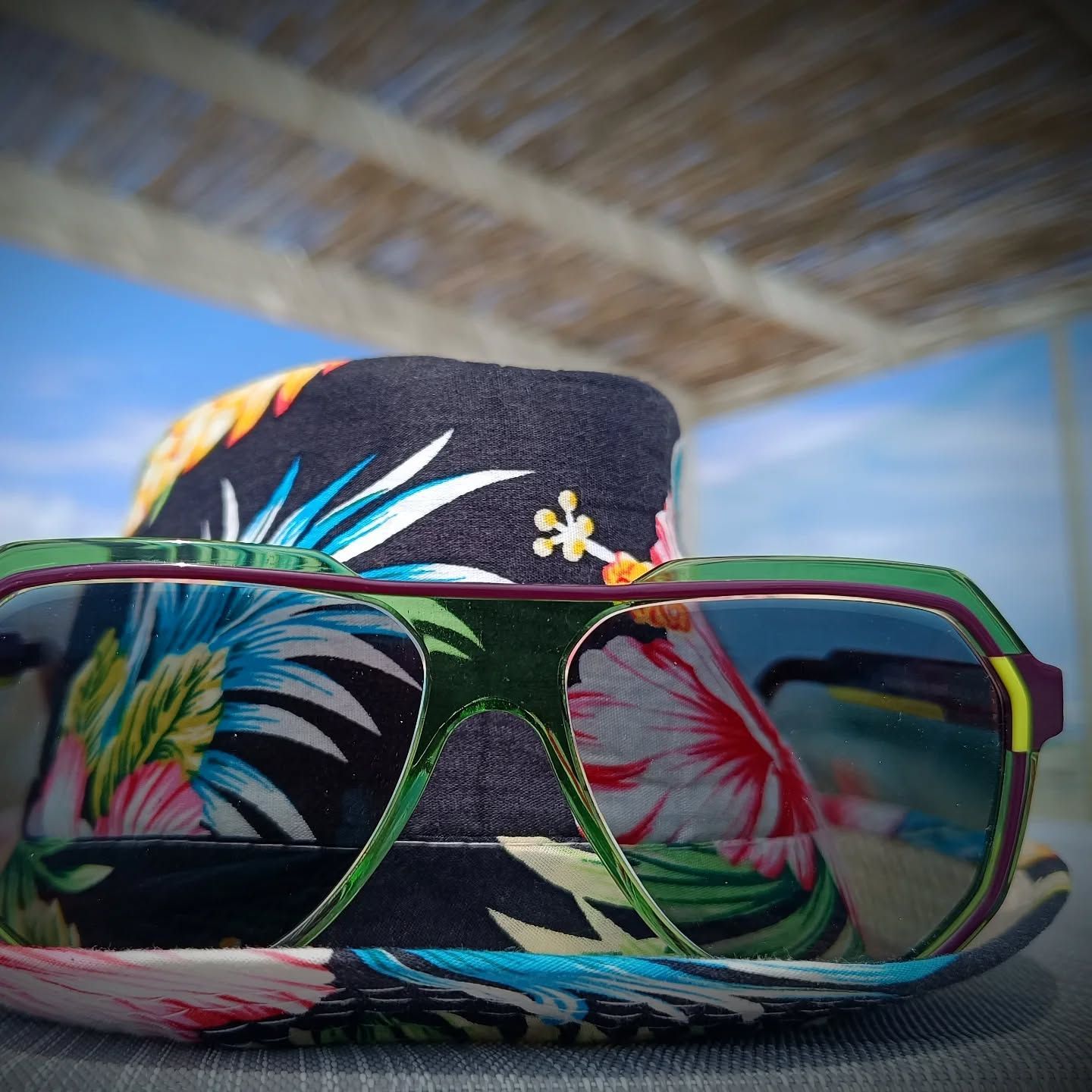 Occhiali da sole floreali colorati su una spiaggia, che riflettono le foglie di palma sotto un cappello da sole.