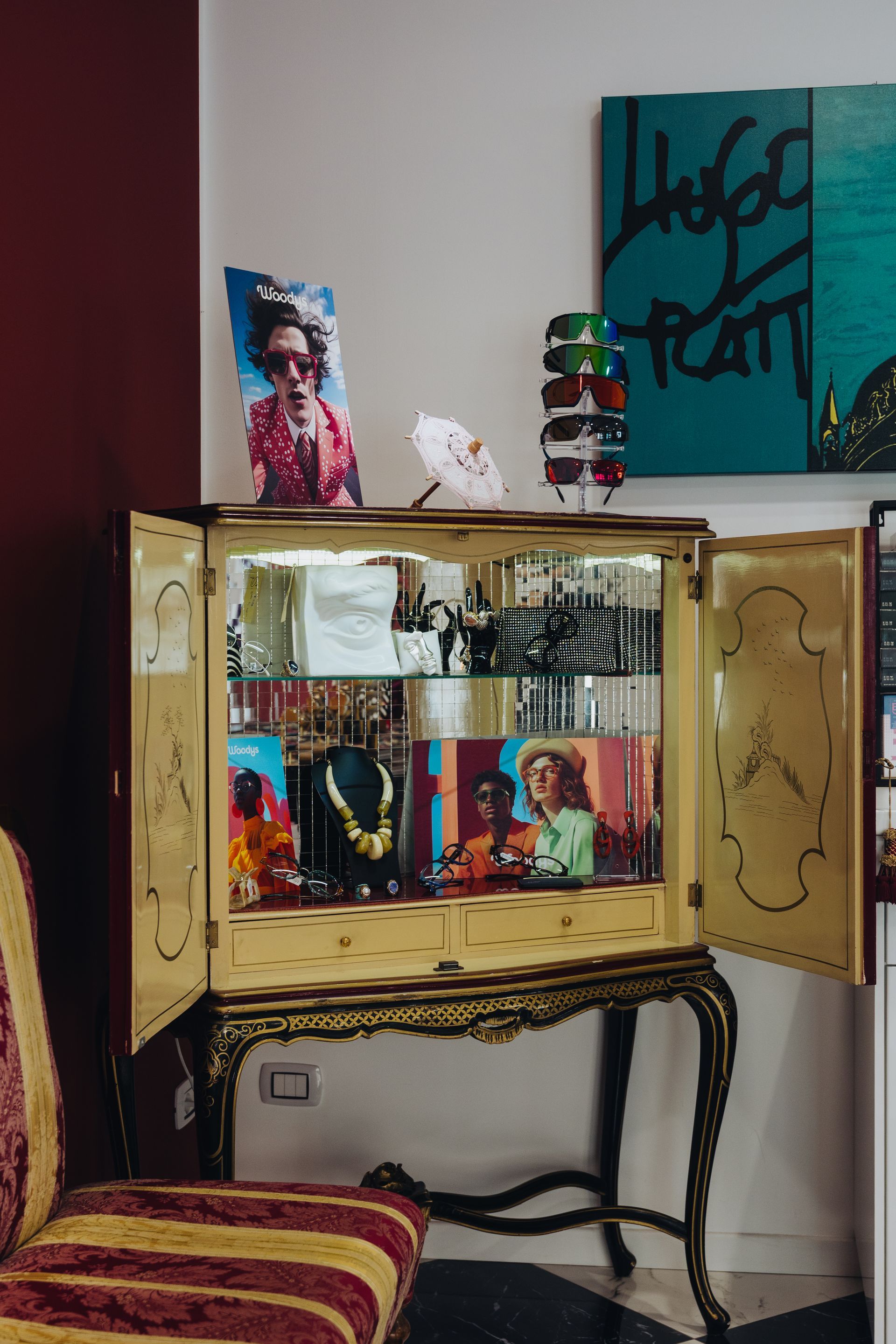 Un piccolo mobiletto vintage con ante in vetro e decorazioni colorate in un angolo, accanto a una sedia a righe e a dei quadri alle pareti.