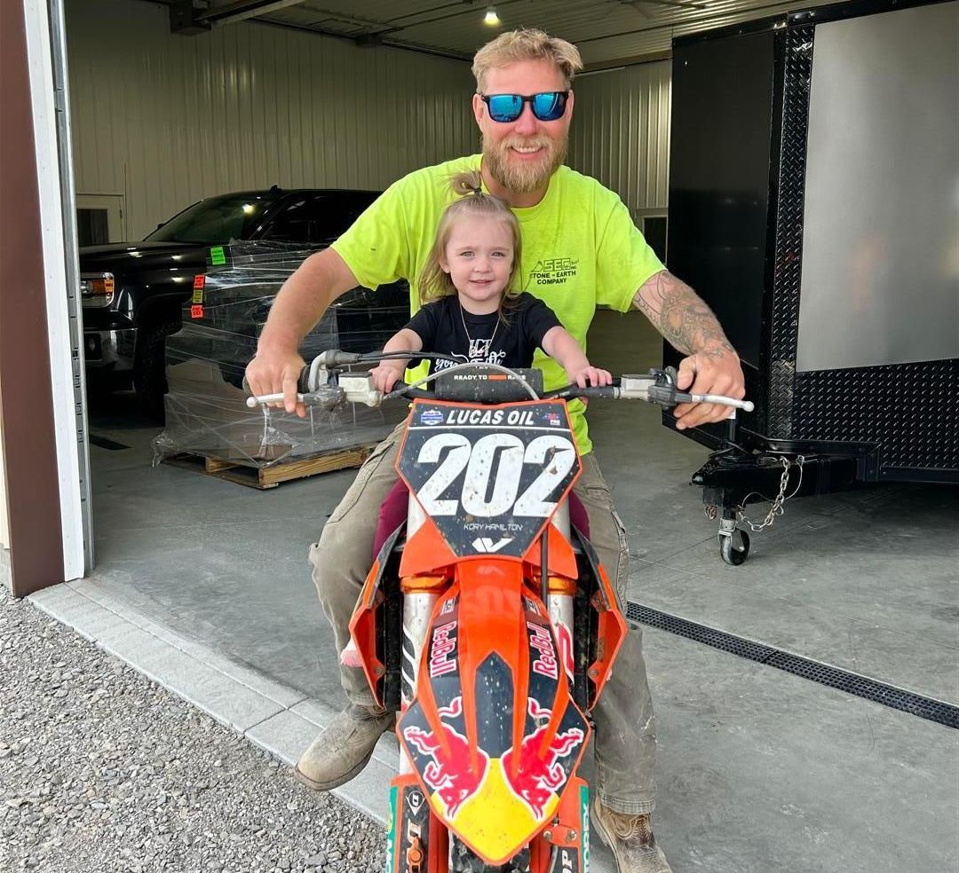 Man and young girl on an orange dirt bike in a garage; man wears sunglasses, yellow shirt; both smiling.