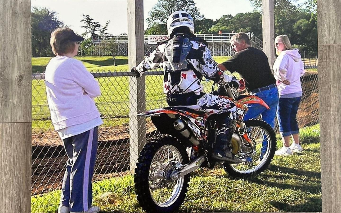 Motocross rider on bike, preparing to ride, with spectators watching at a dirt track.
