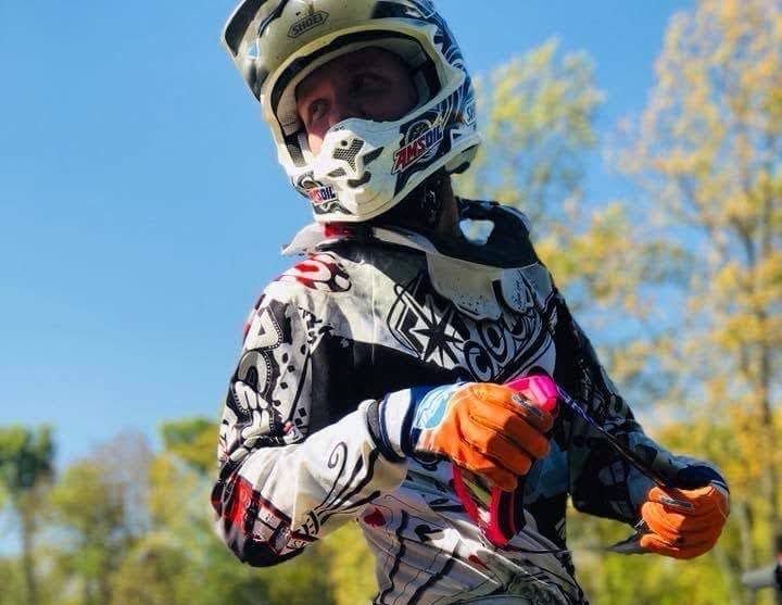 Person in motorcycle helmet and gear, outdoors, holding item, with trees and blue sky in background.