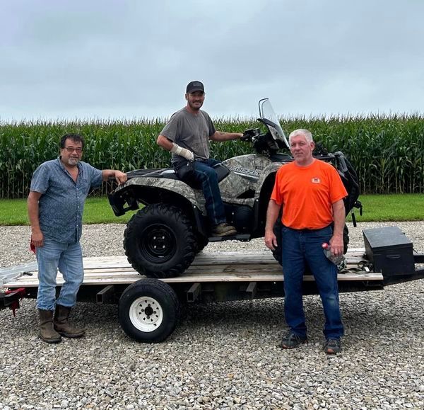 Three men with an ATV on a trailer in front of a cornfield.