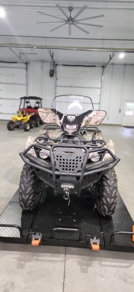 An ATV with a camo print, parked on a black trailer in a garage, a yellow vehicle in the background.