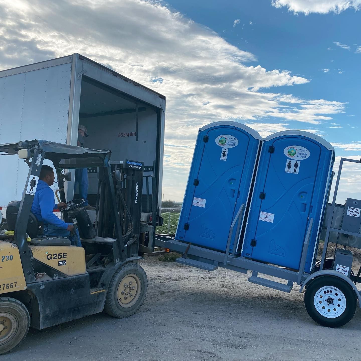 A man is driving a forklift pulling two blue portable toilets on a trailer.