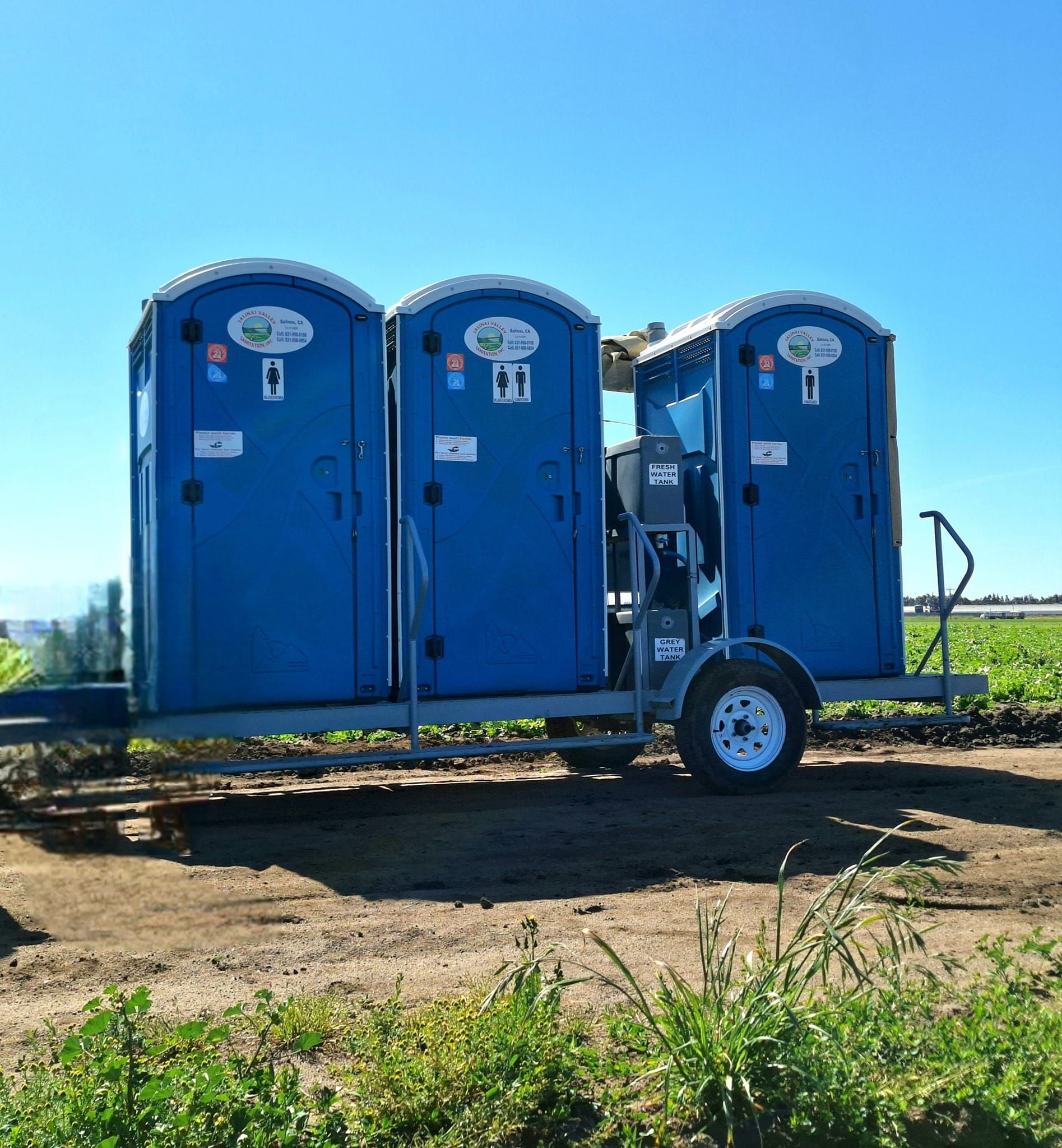 Three blue portable toilets are on a trailer in a field.