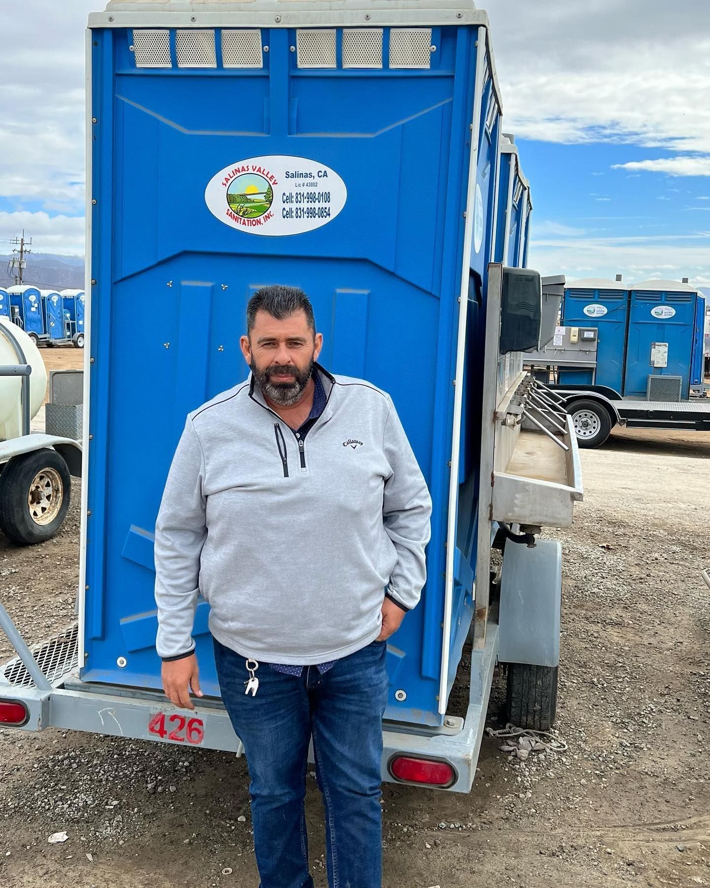A man is standing in front of a blue portable toilet on a trailer.
