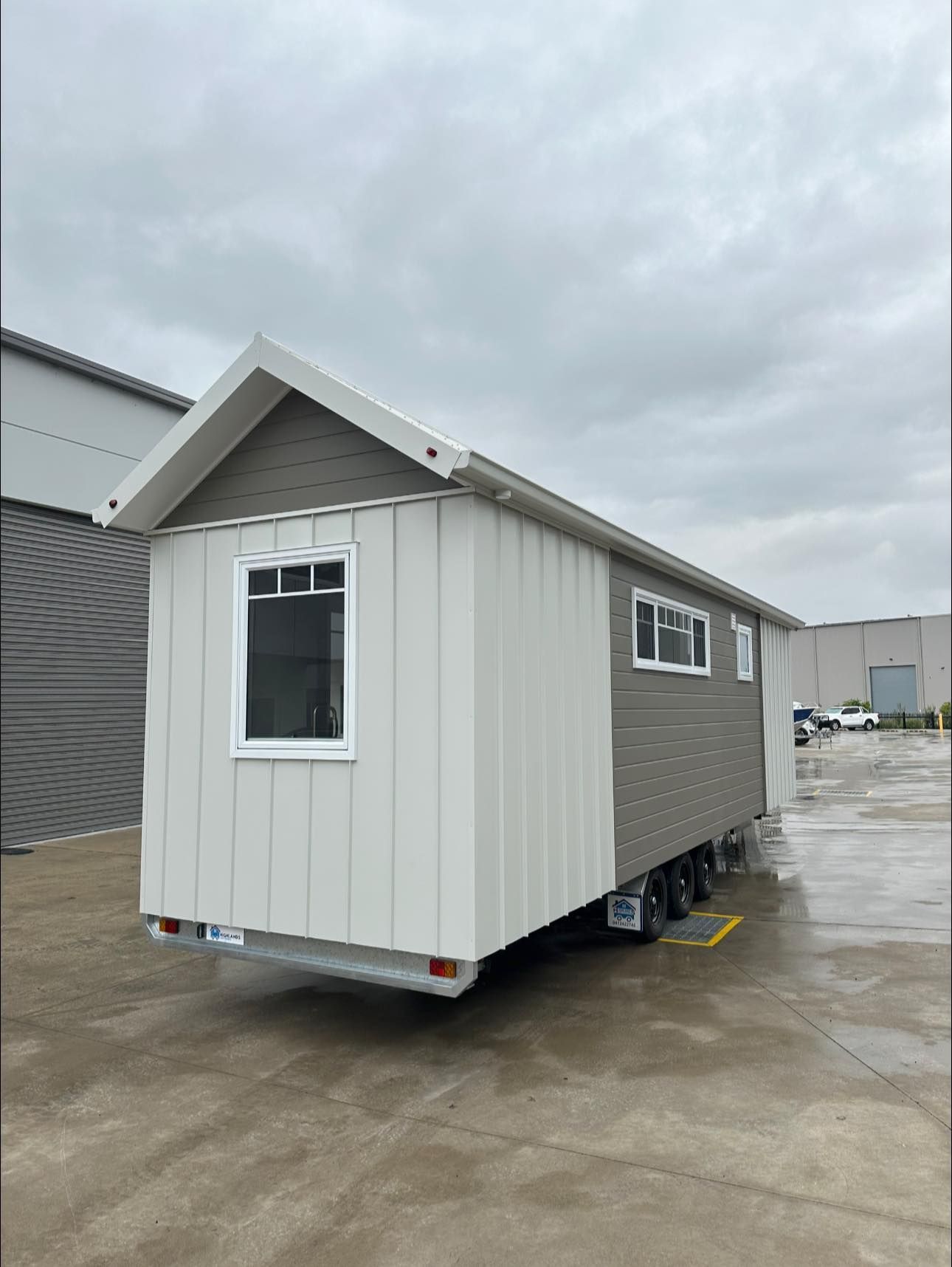 Tiny house on wheels with gray and white siding, parked outside a building on a cloudy day.