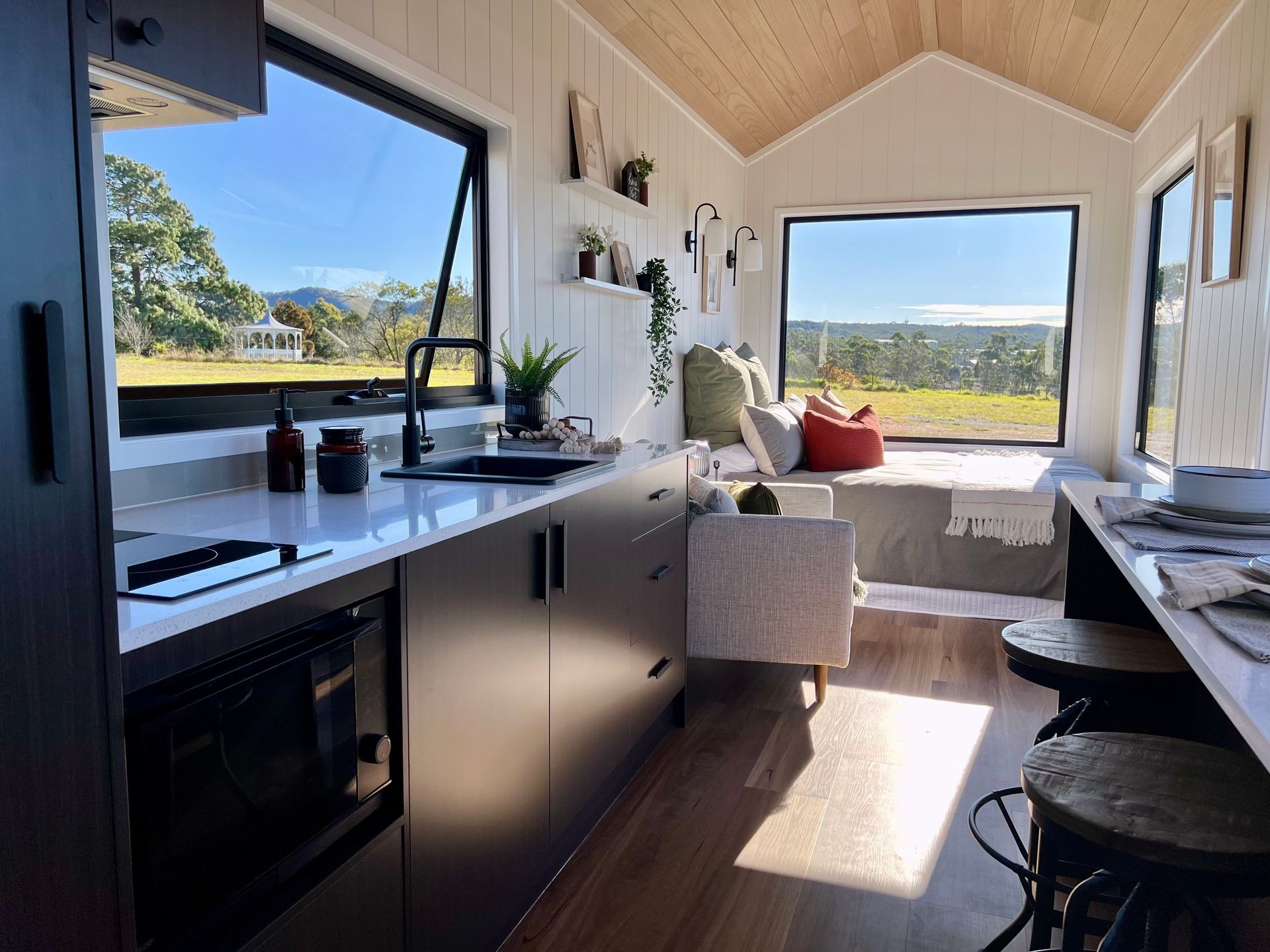 Interior of a tiny home kitchen with a large window. Black cabinets, white countertops, wooden ceiling and views of a field.