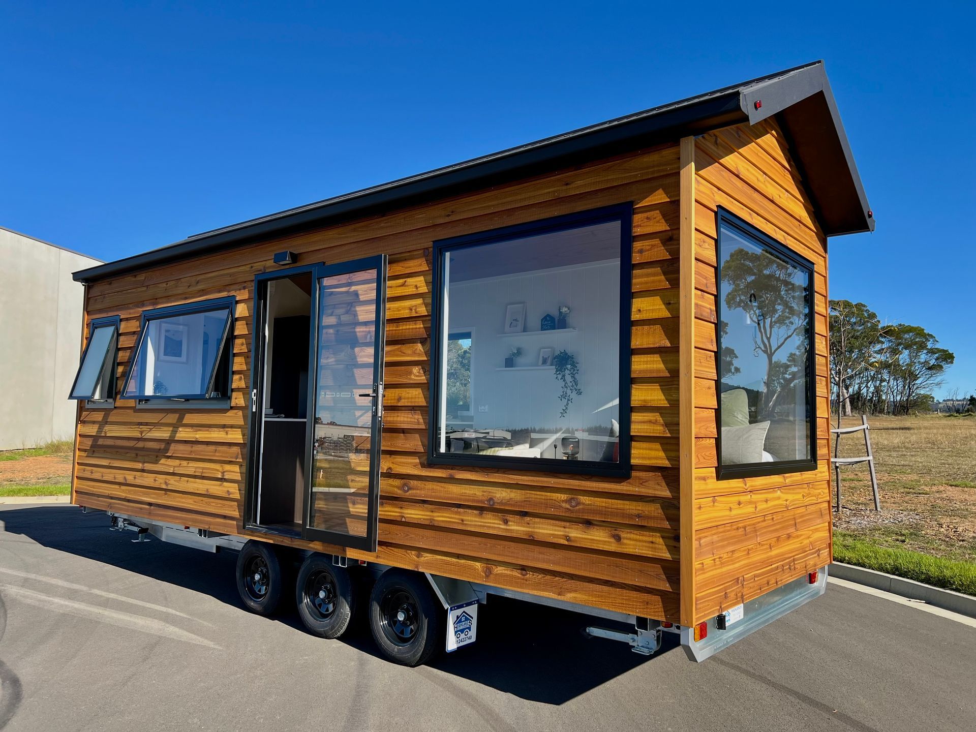 Tiny wooden house on wheels with large windows, parked on a sunny day.