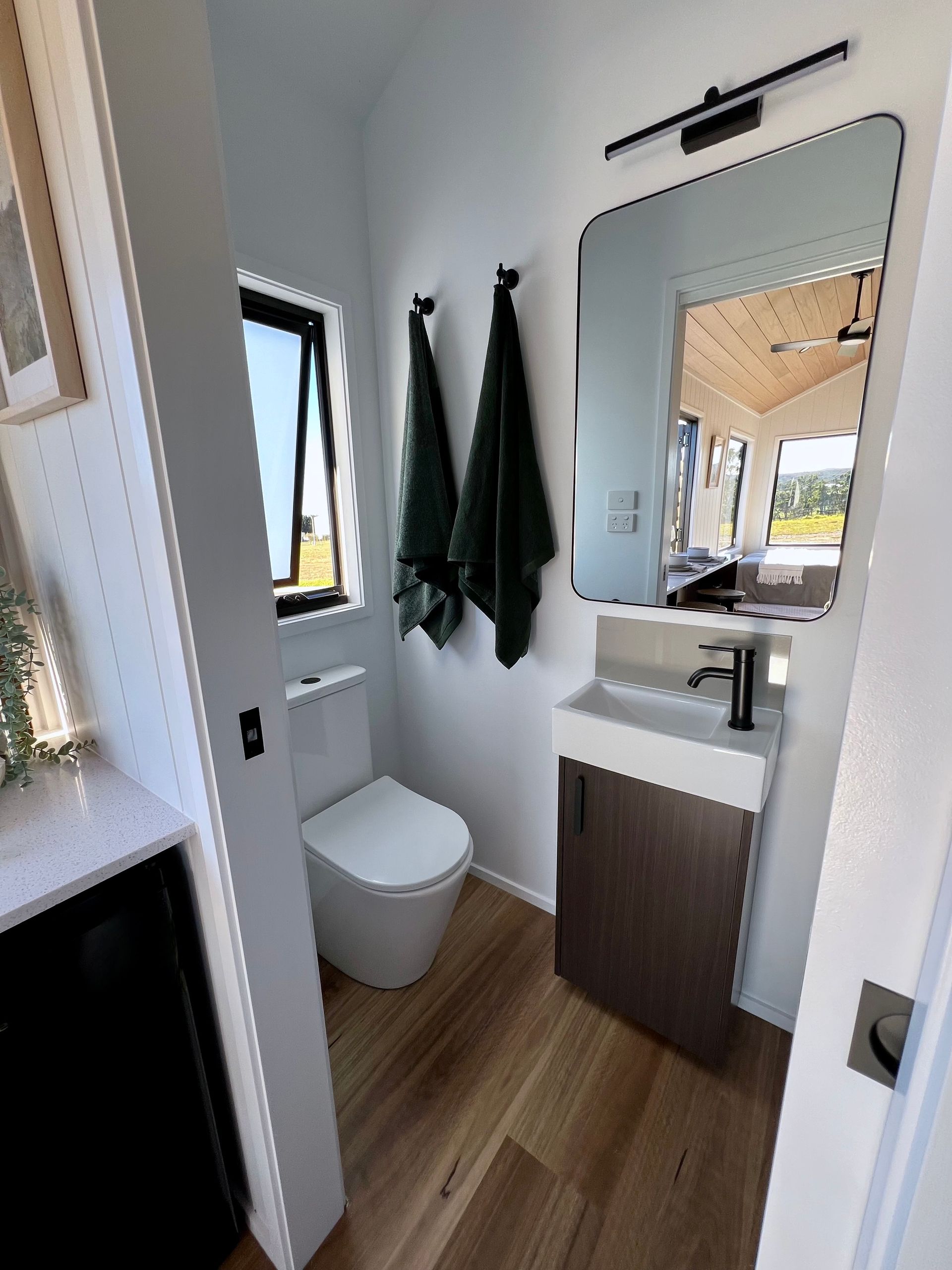 Small bathroom with white toilet, sink, and dark wood cabinet. Black fixtures, green towels, and wooden floor.