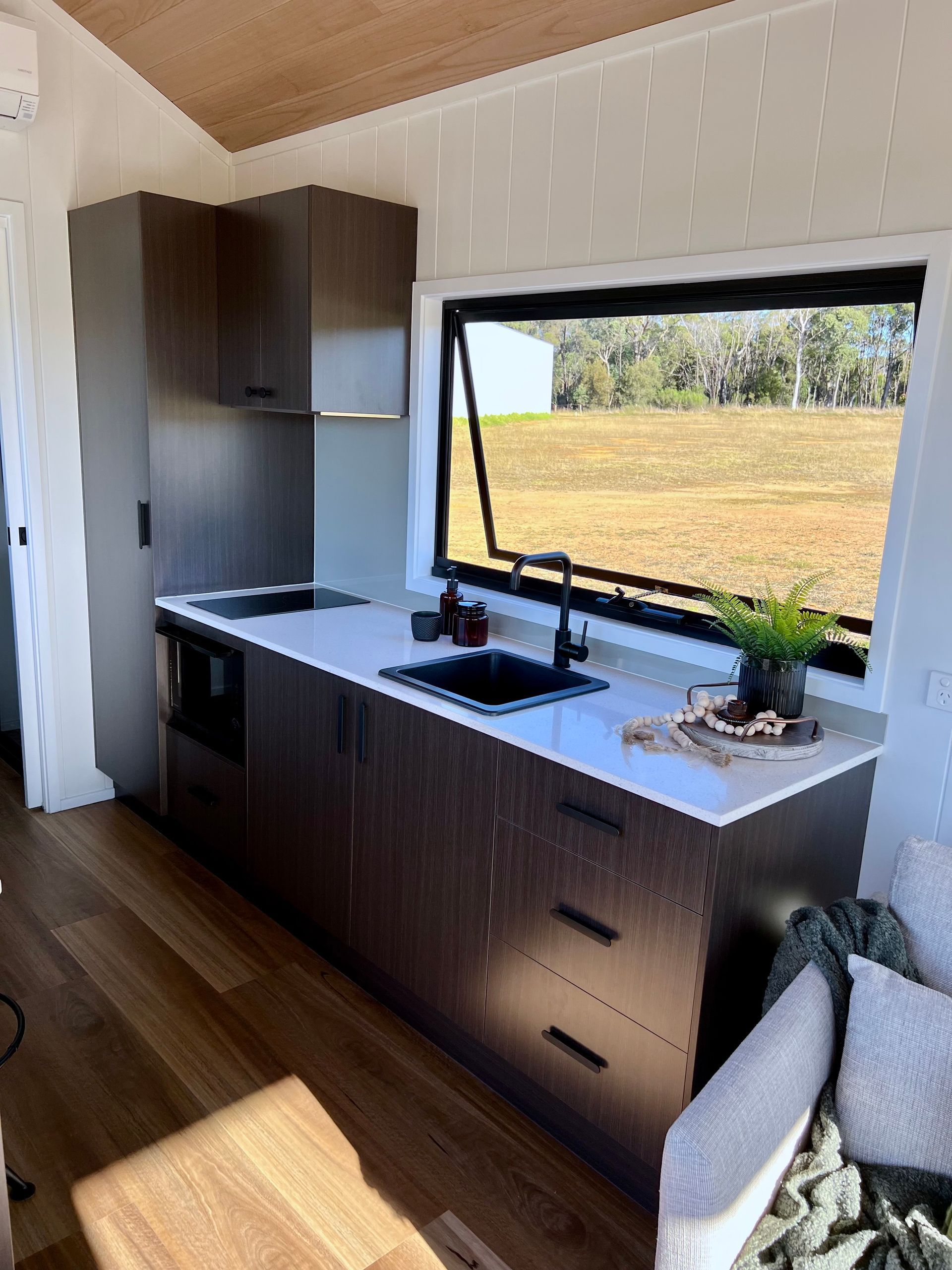 Small kitchen with dark cabinets, white countertop, and open window overlooking a field.