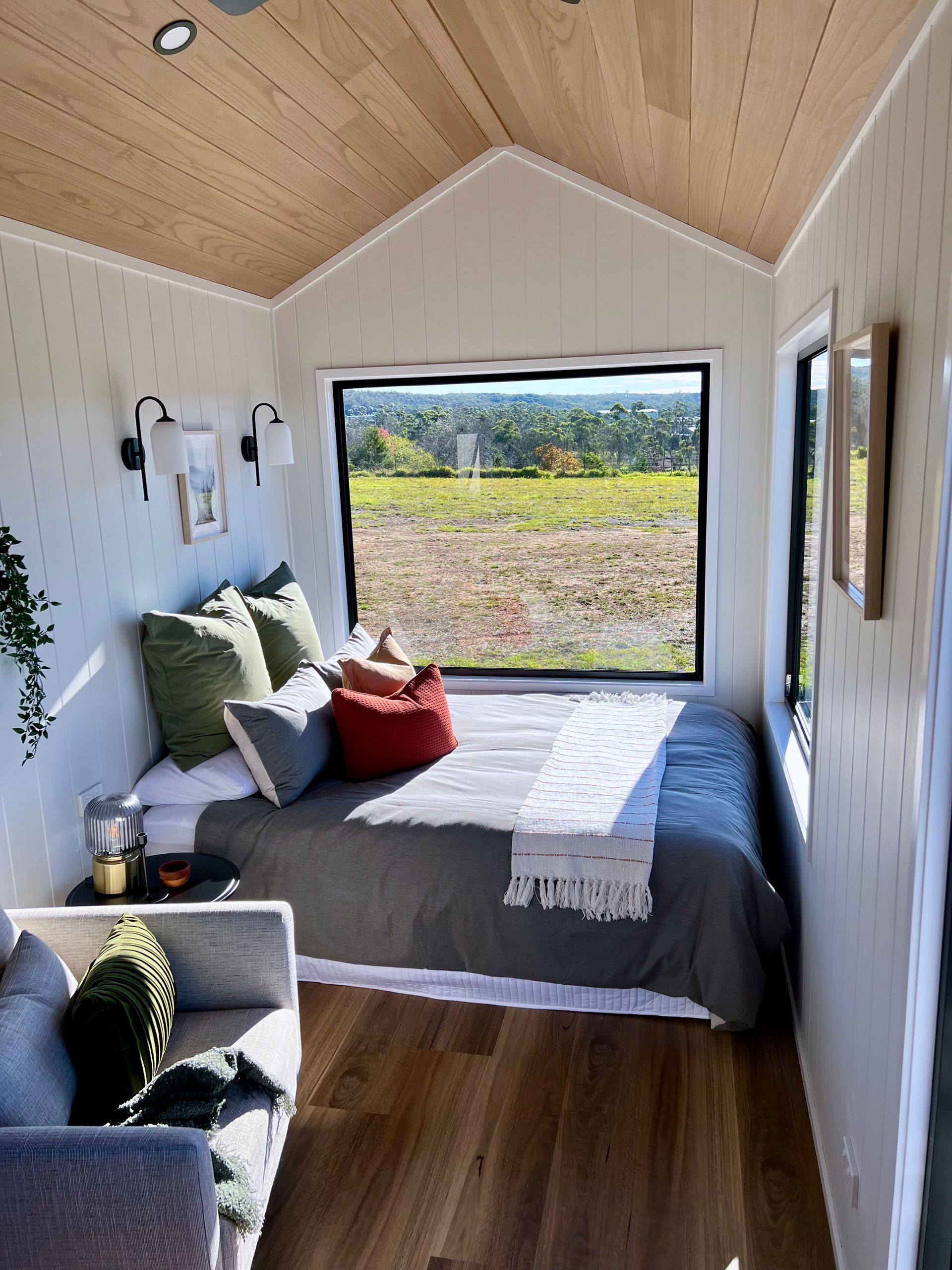 Cozy tiny bedroom with bed, large window overlooking field, light wood ceiling and floor.